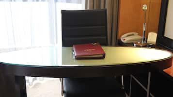 Ett hotellrum med ett bord med glasskiva, en röd bok, en svart stol, en vit telefon och en spegel.