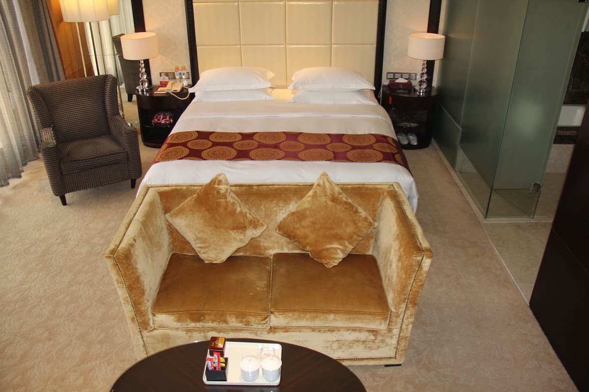 Quarto de hotel com uma cama grande, uma poltrona marrom, uma mesinha com um porta-objetos e uma área de chuveiro com vidro.