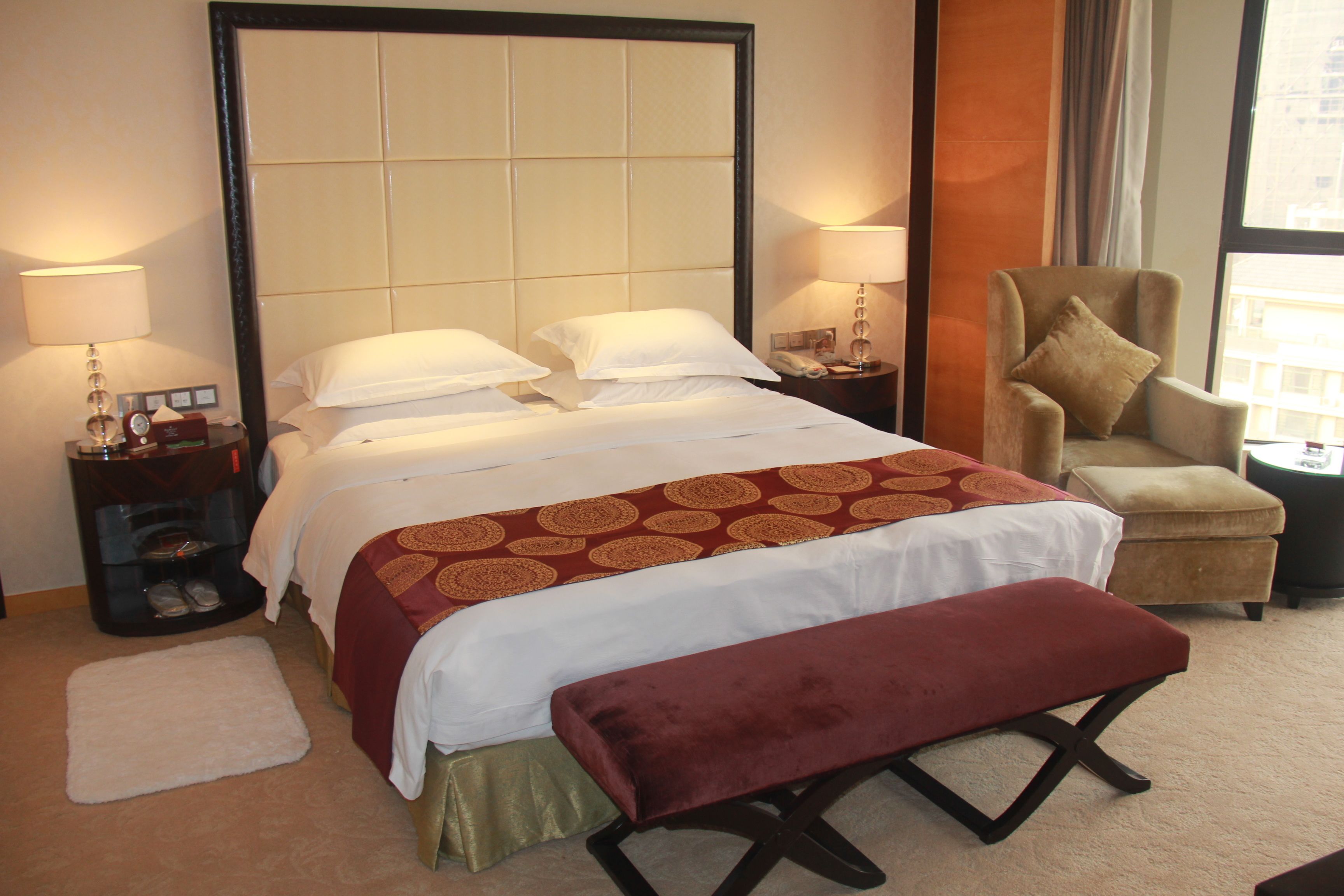 A hotel room with a large bed, two bedside tables, a chair, a bench, and a window with a view.