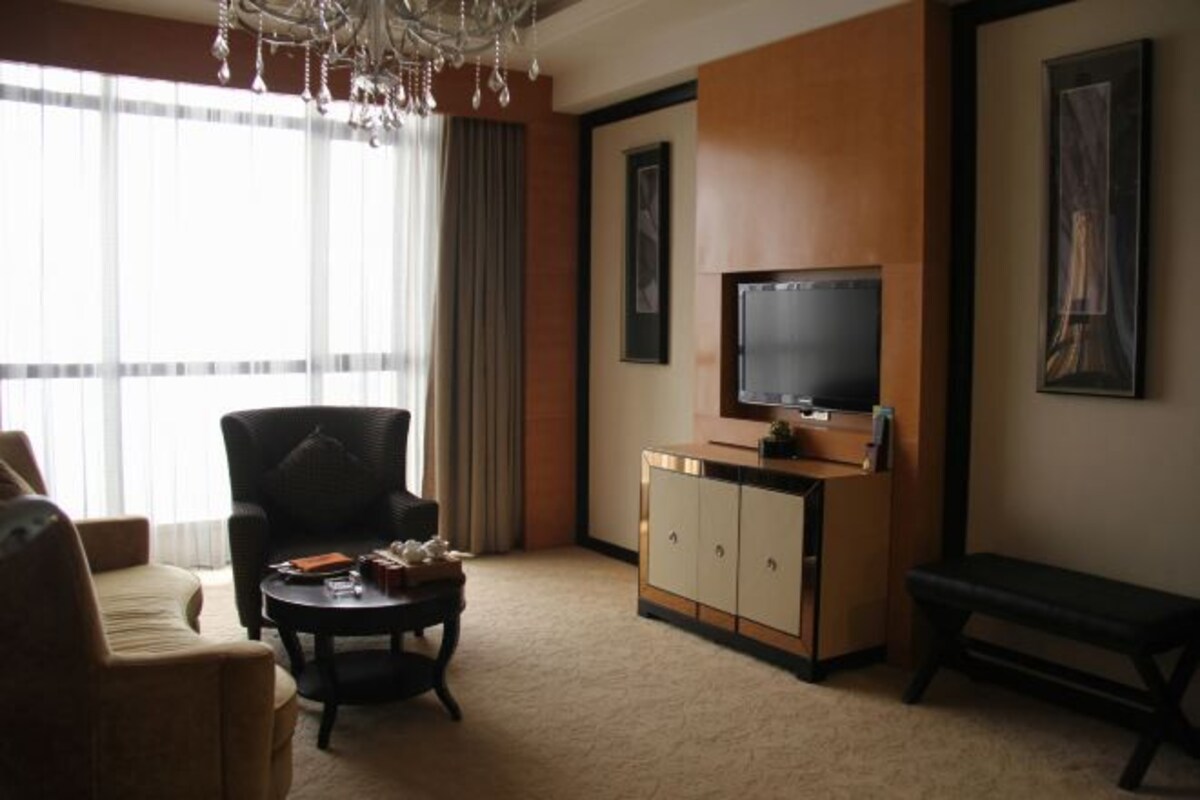 Habitación de hotel con un ventanal grande, televisión de pantalla plana, sofá, sillón, mesa de centro y un banco.