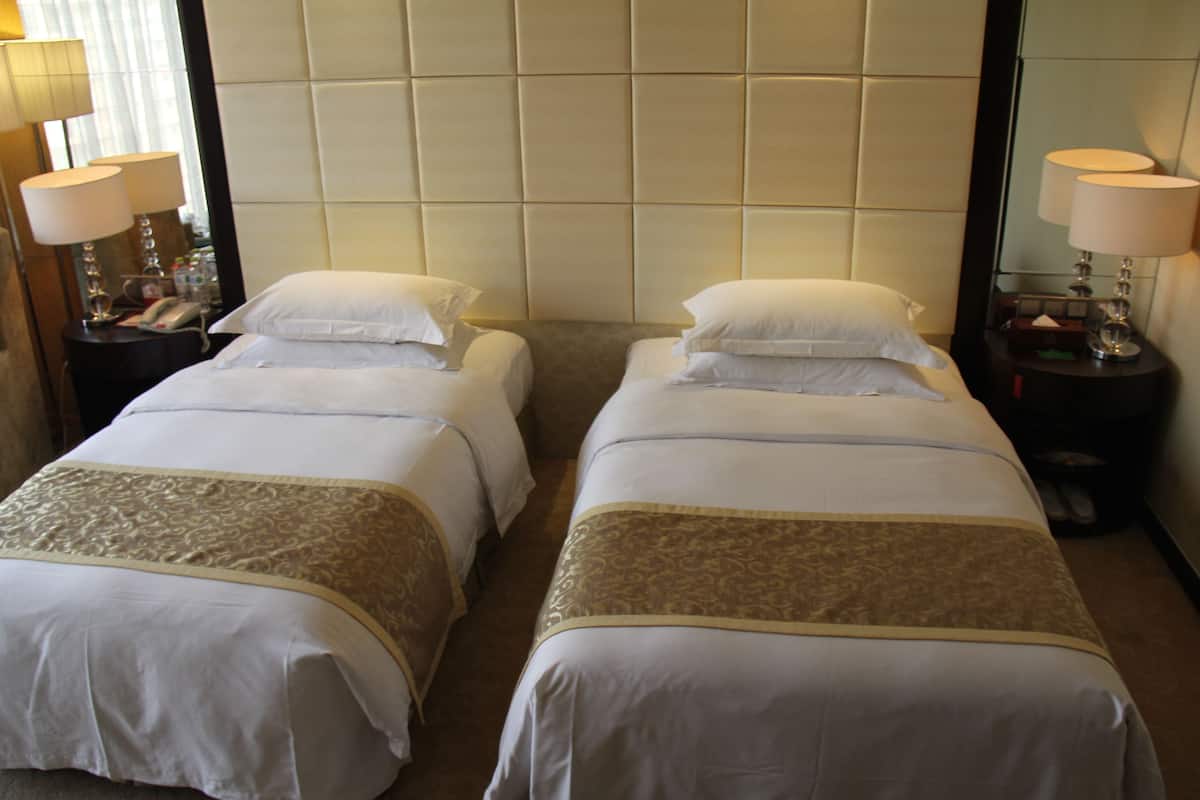 Quarto de hotel com duas camas individuais, cada uma com colcha branca e manta com padrão dourado. Há mesas de cabeceira com luminárias, um espelho e um telefone.