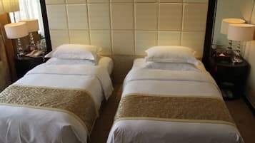 Een hotelkamer met twee enkele bedden, elk met een witte sprei en een gouden sierkleed. Er zijn nachtkastjes met lampen, een spiegel en een telefoon.