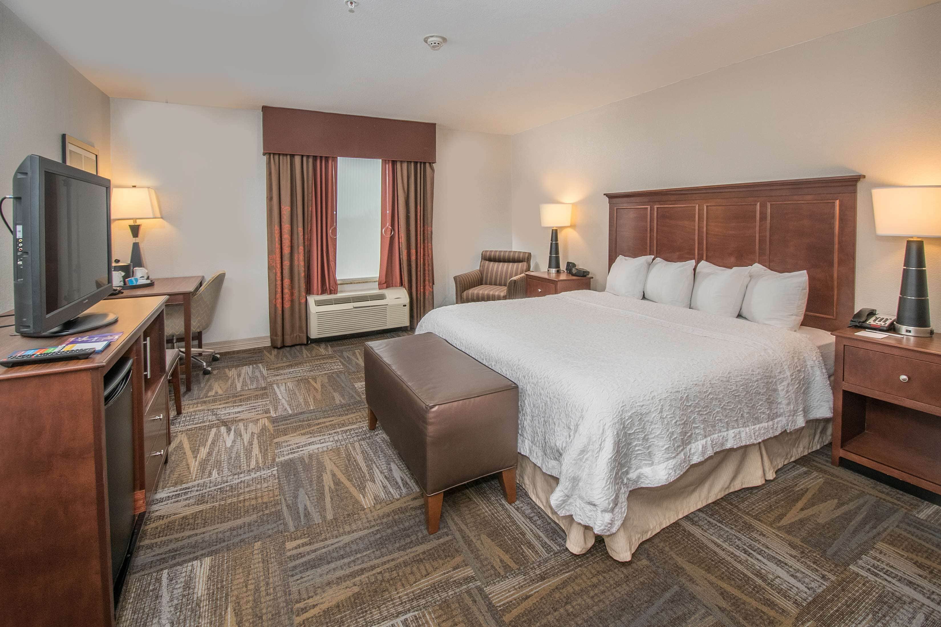 A hotel room with a large bed, a desk with a television, a chair, a bedside table with a lamp, and a carpeted floor.