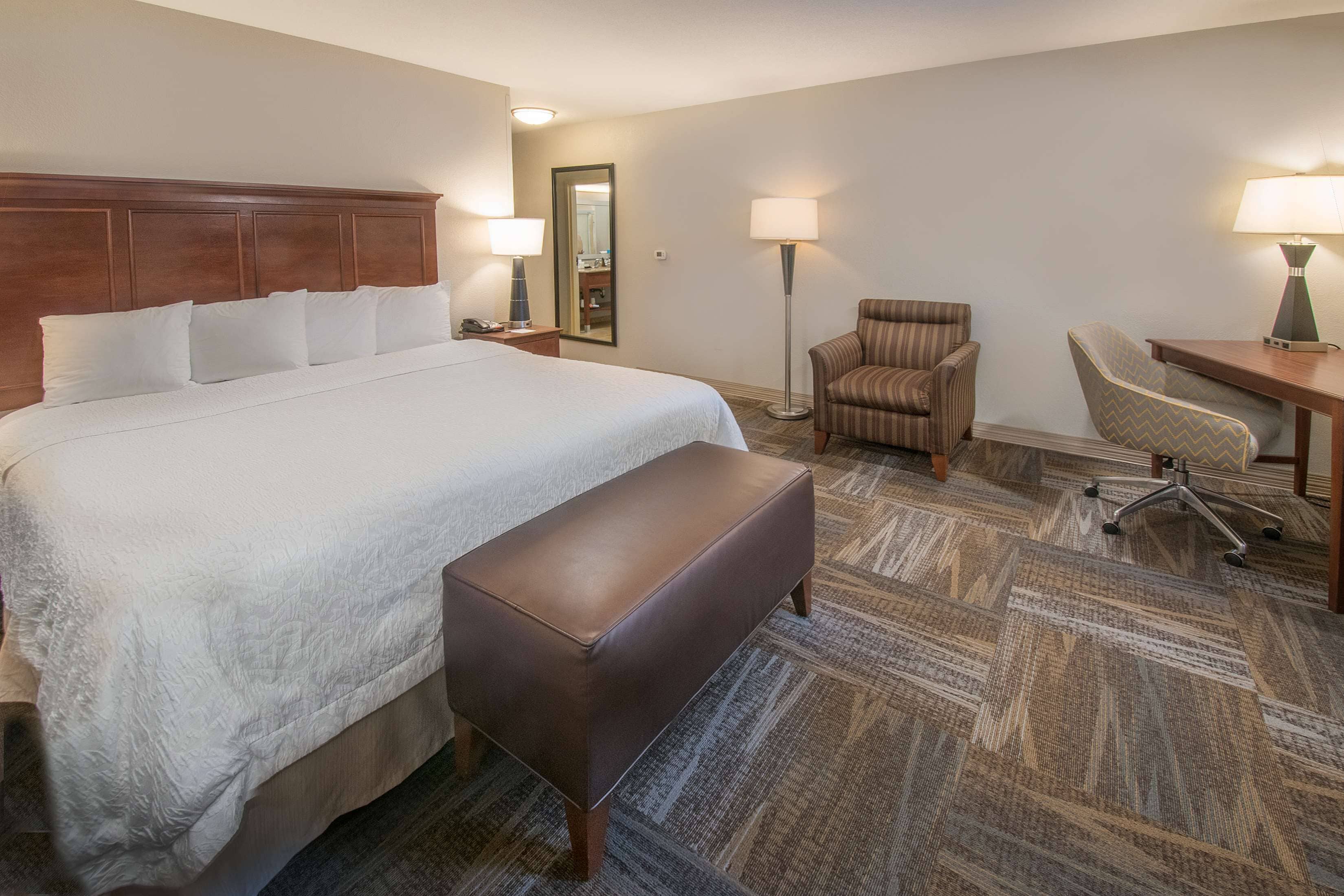 A hotel room with a large bed, a desk, two chairs, a bench, and two lamps.