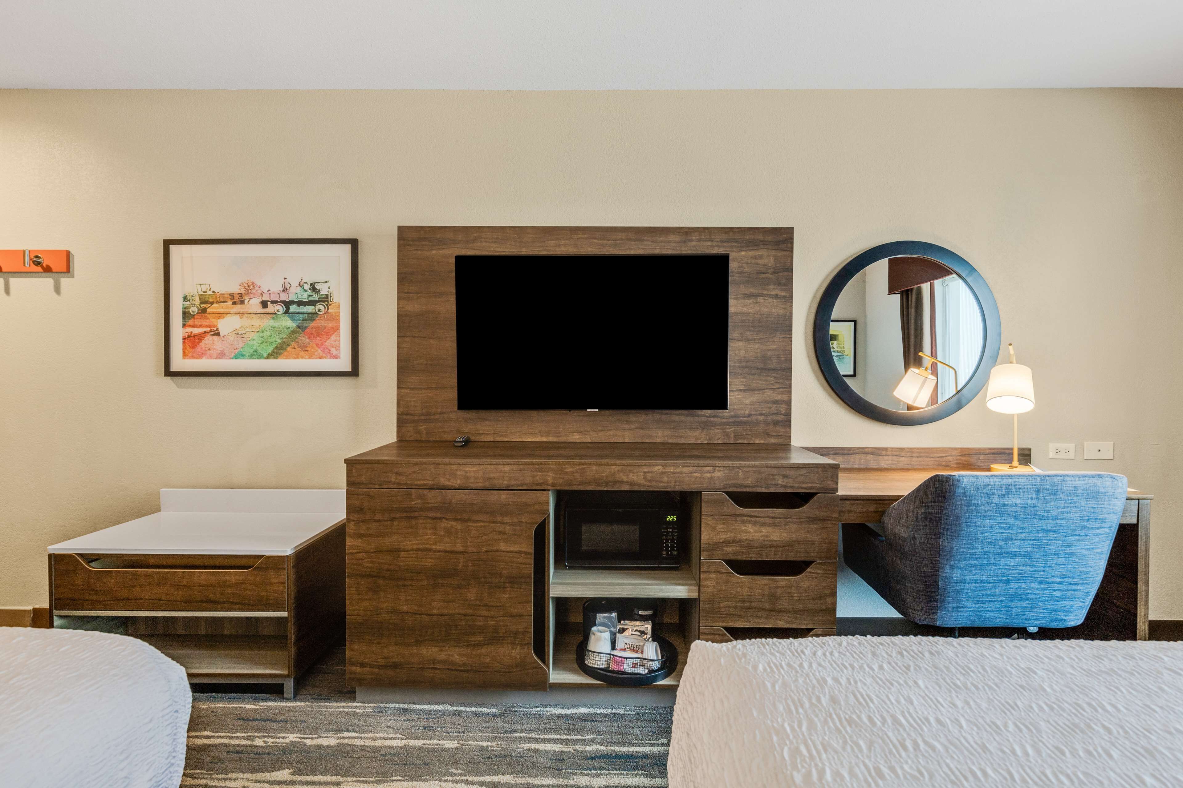 Une chambre d’hôtel dotée d’un téléviseur à écran plat fixé au-dessus d’un bureau en bois, d’un miroir rond et d’un lit recouvert d’une couette blanche.