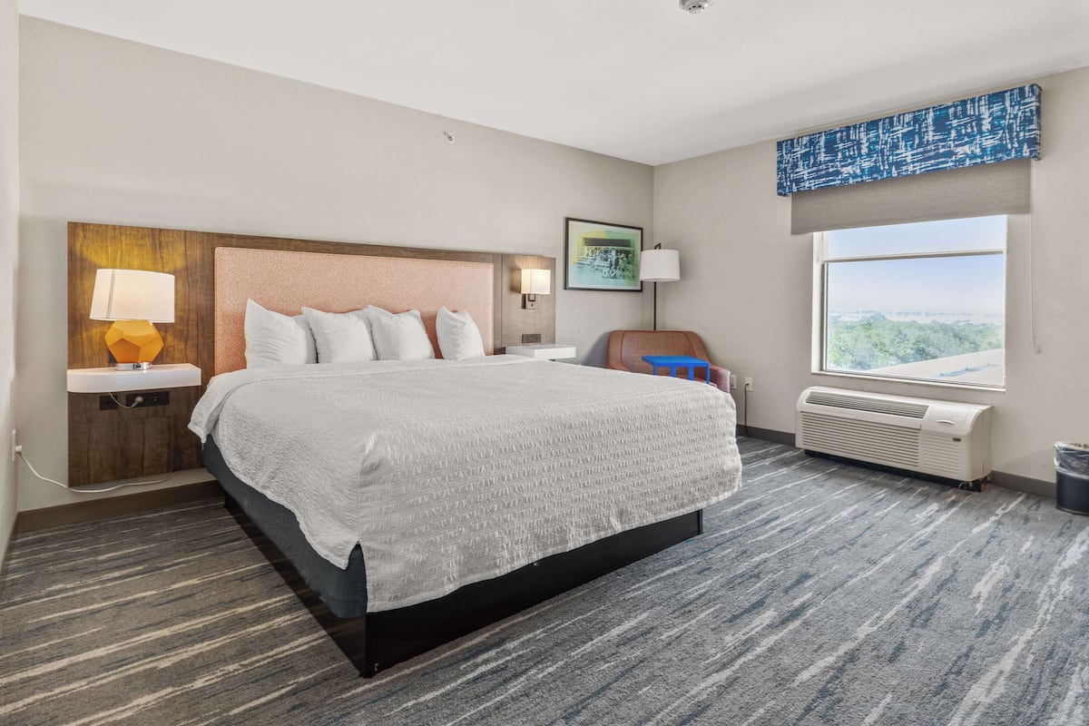 Habitación de hotel con una cama grande, una mesita de noche, una lámpara, una silla, un ventanal con vistas y un aire acondicionado.