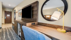 Habitación de hotel con un escritorio de madera, un televisor de pantalla plana, un espejo redondo y una lámpara.