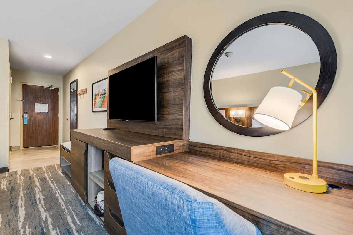 Habitación de hotel con un escritorio de madera, un televisor de pantalla plana, un espejo redondo y una lámpara.