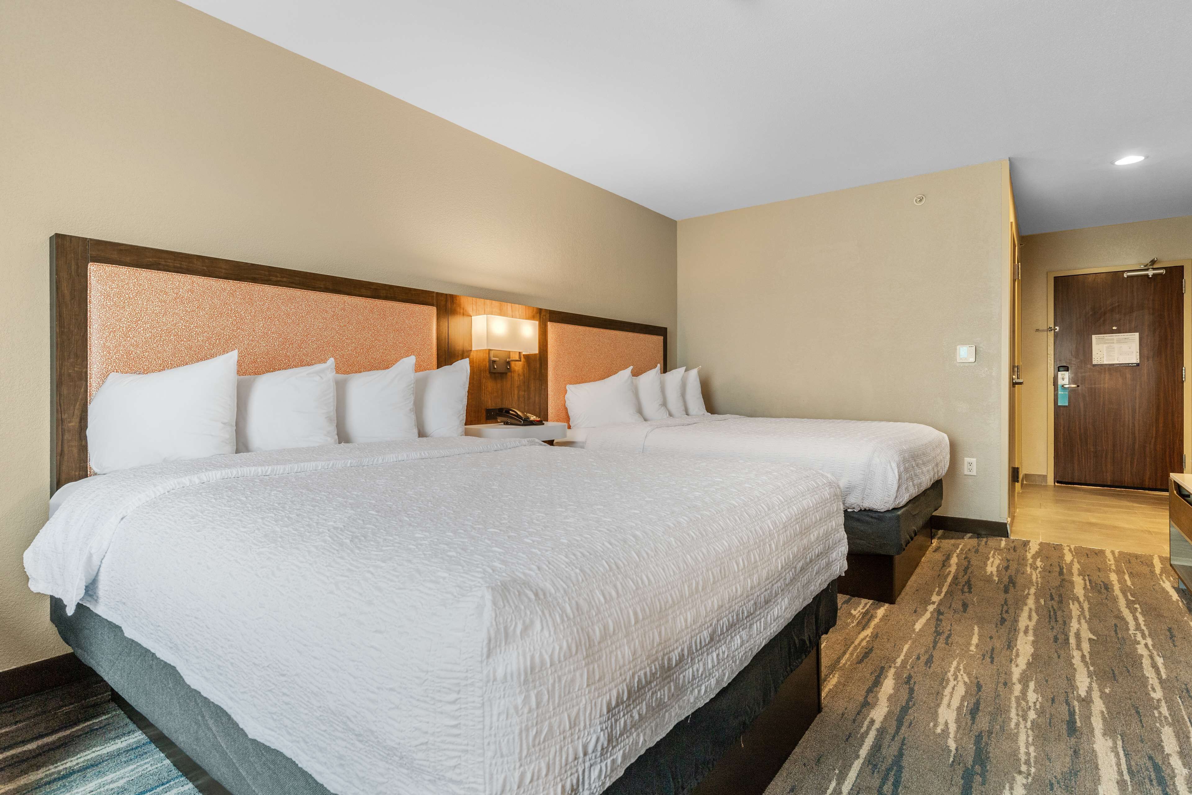 Une chambre d’hôtel avec deux lits, une tête de lit dotée d’un luminaire, une porte en bois et un sol recouvert de moquette.