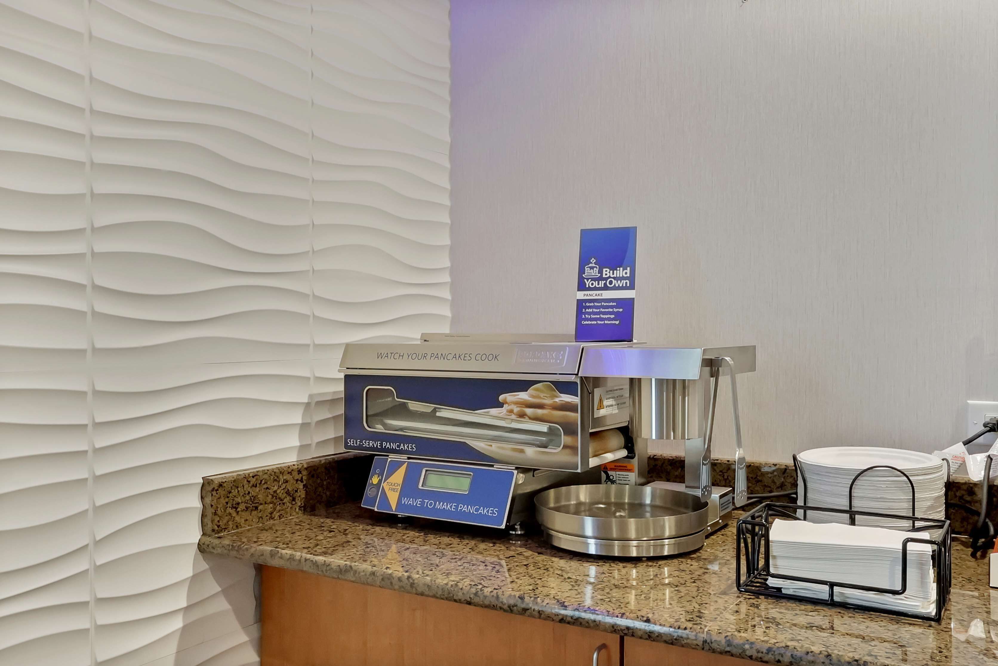 A self-serve pancake station with a digital interface, a metal bowl, and a stack of plates.