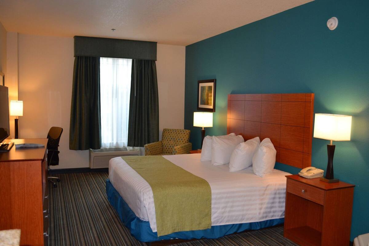 A hotel room with a bed, a desk, a chair, a desk lamp, a bedside table with a lamp, a telephone, and a window with curtains.