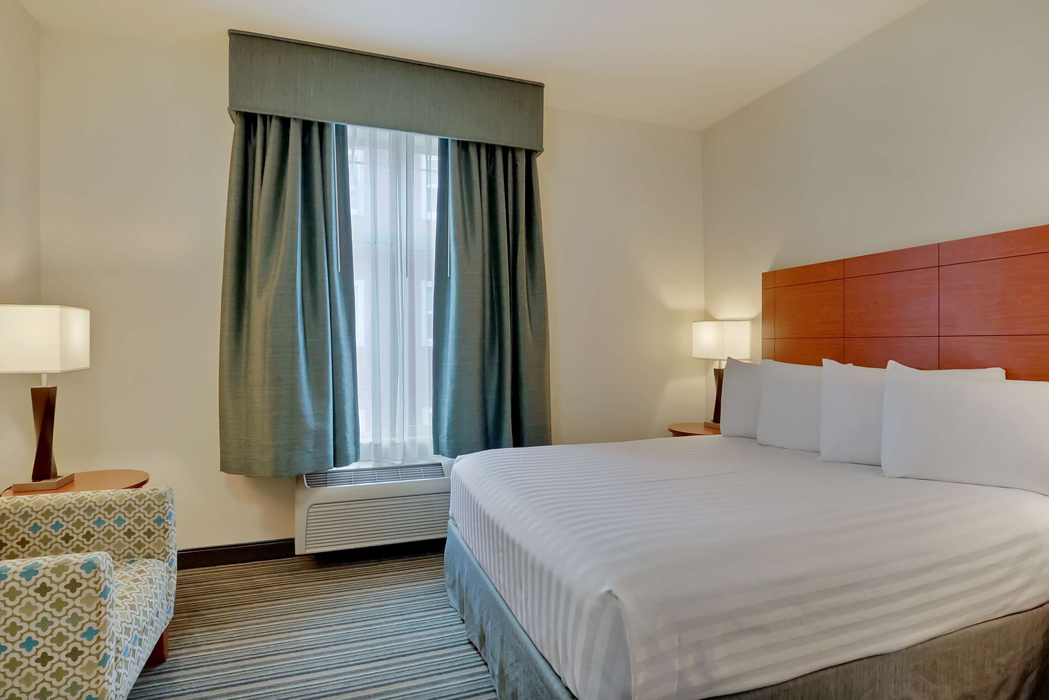 Ein Hotelzimmer mit einem großen Bett, einem Nachttisch mit Lampe, einem gemusterten Sessel, einem Fenster mit Vorhängen und einer Klimaanlage.