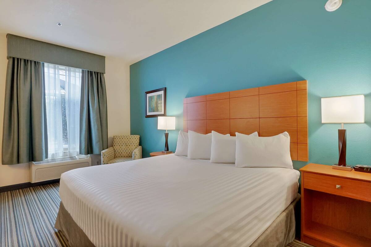 A hotel room with a large bed, a wooden headboard, two bedside lamps, a chair, a picture on the wall, and a window with curtains.