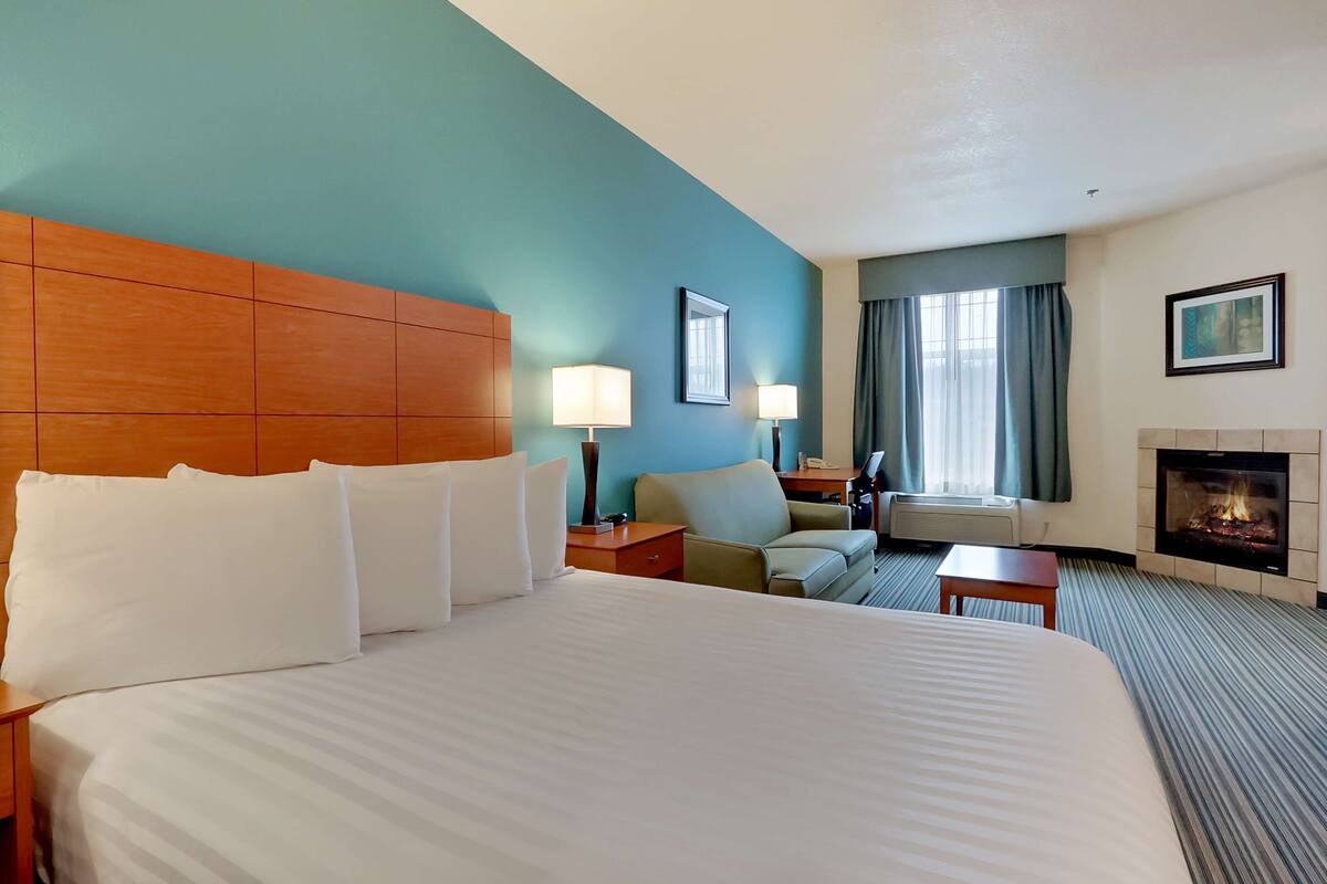 A hotel room with a large bed, two bedside tables with lamps, a sofa, a small table, a fireplace, and a window with curtains.