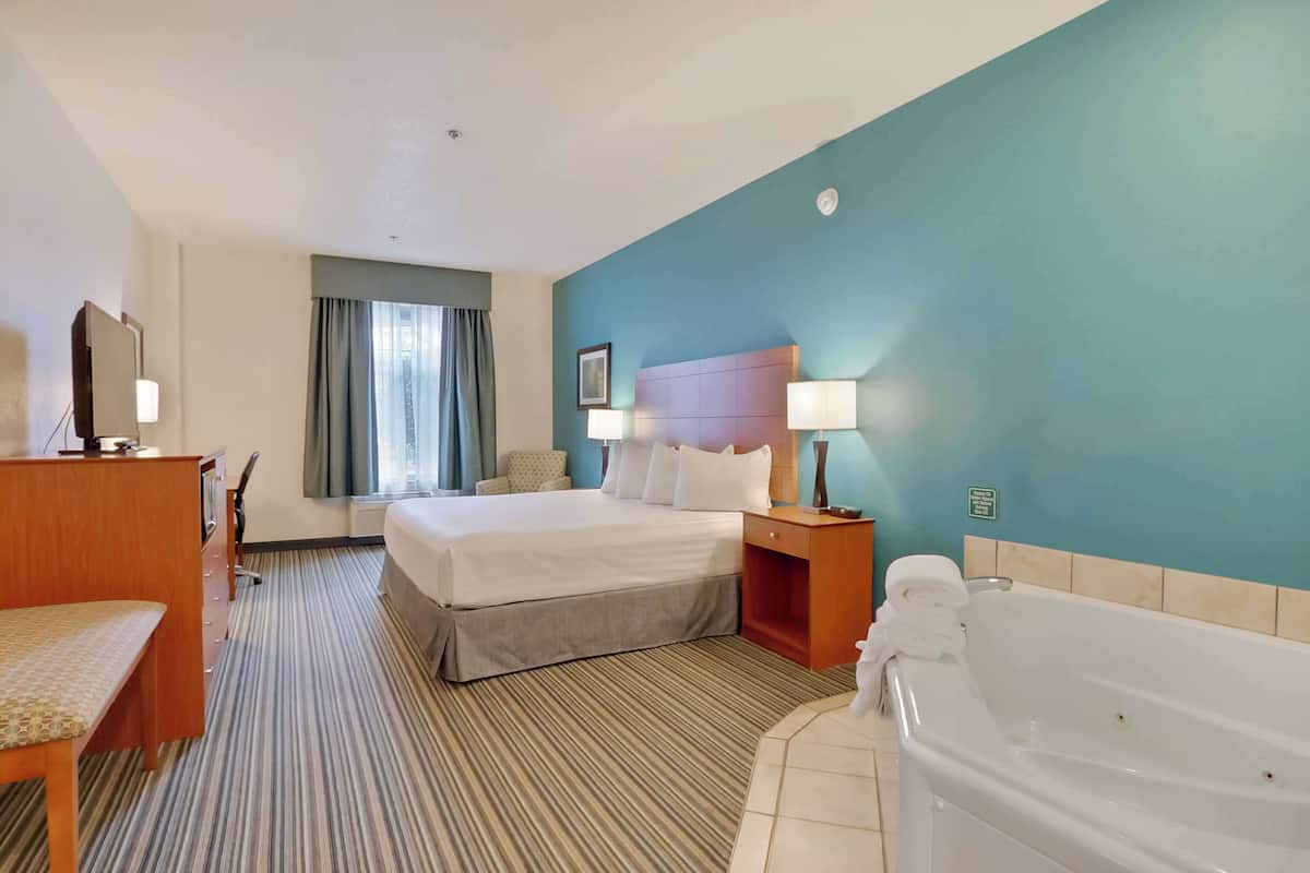 A hotel room with a bed, a desk, a chair, a desk lamp, a nightstand, a lamp, a bathroom with a bathtub, and a window with curtains.