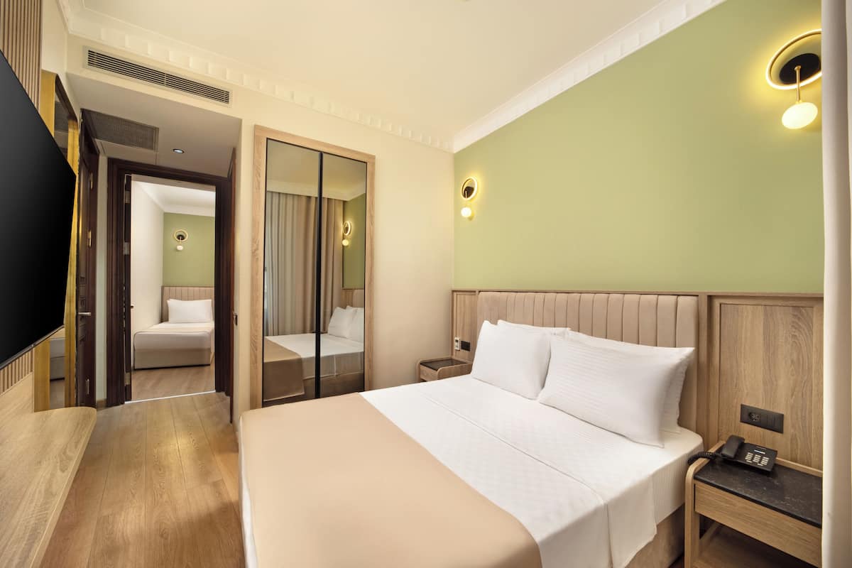 A hotel room with a large bed, a bedside table with a phone, a mirror, and a door leading to another room.