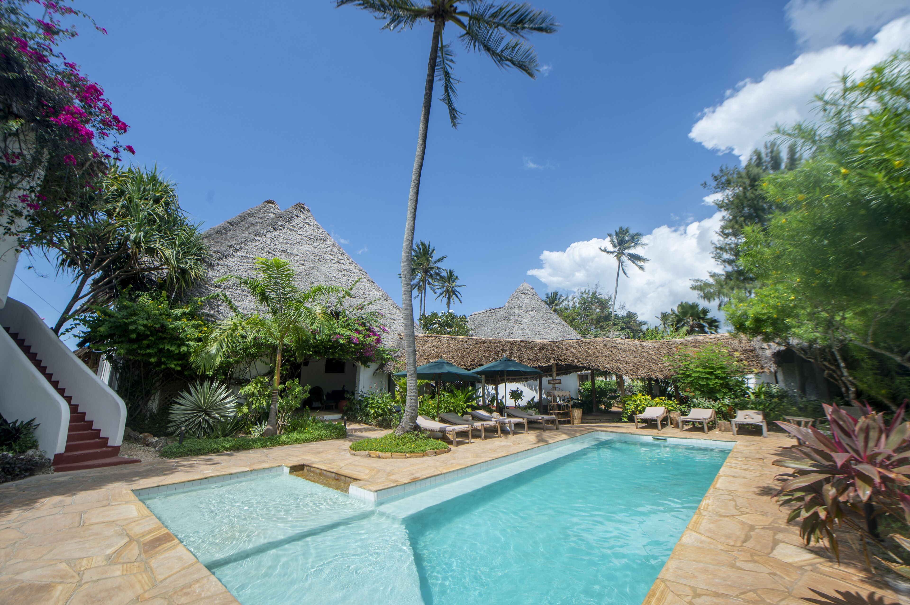 A pool area with lounge chairs, a staircase, and thatched-roof structures.