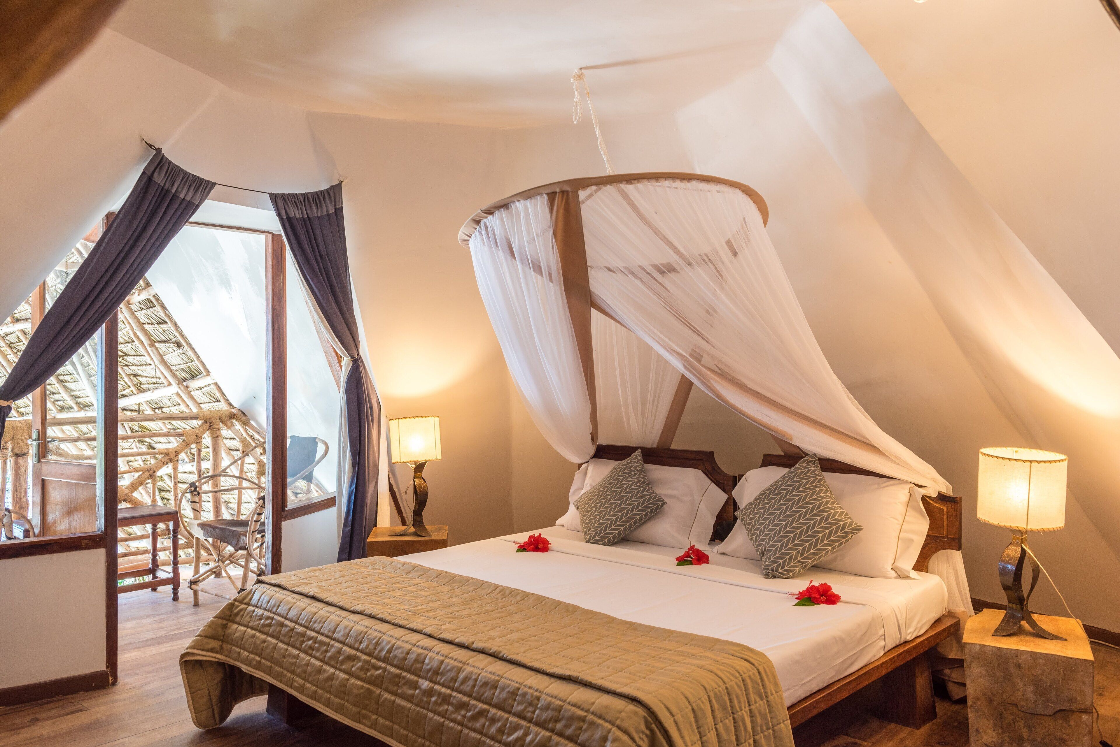 Quarto com uma cama de dossel, cabeceira de madeira e cortina. Há uma cadeira e uma mesa perto da janela, e um abajur na mesinha de cabeceira.