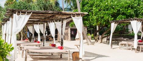 A beachside cabana area with wooden beds and white canopies, surrounded by palm trees and greenery.