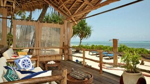 Une terrasse en bois surmontée d’un toit de chaume, donnant sur une plage. On y trouve des chaises longues et une petite table agrémentée d’un vase décoratif.