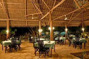 An outdoor dining area with tables and chairs under a thatched roof, surrounded by stone flooring and greenery.