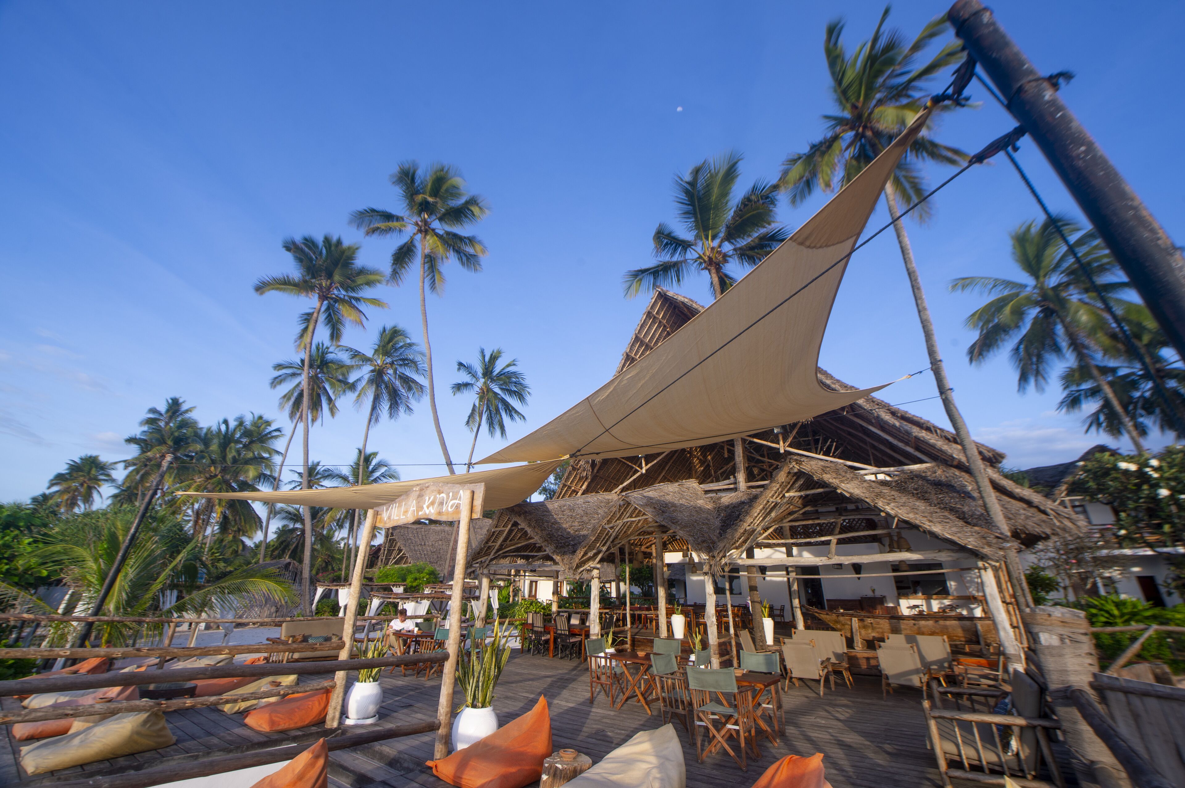 A beachside restaurant with outdoor seating, palm trees, and a thatched roof structure.