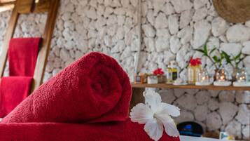 Una toalla roja con una flor blanca sobre una cama, una pared de piedra y un estante con artículos de aseo.