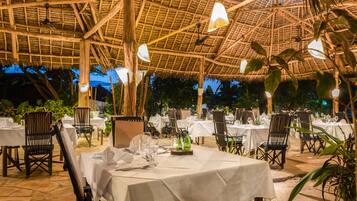 An outdoor dining area with tables set for guests, surrounded by wooden chairs and a thatched roof structure.