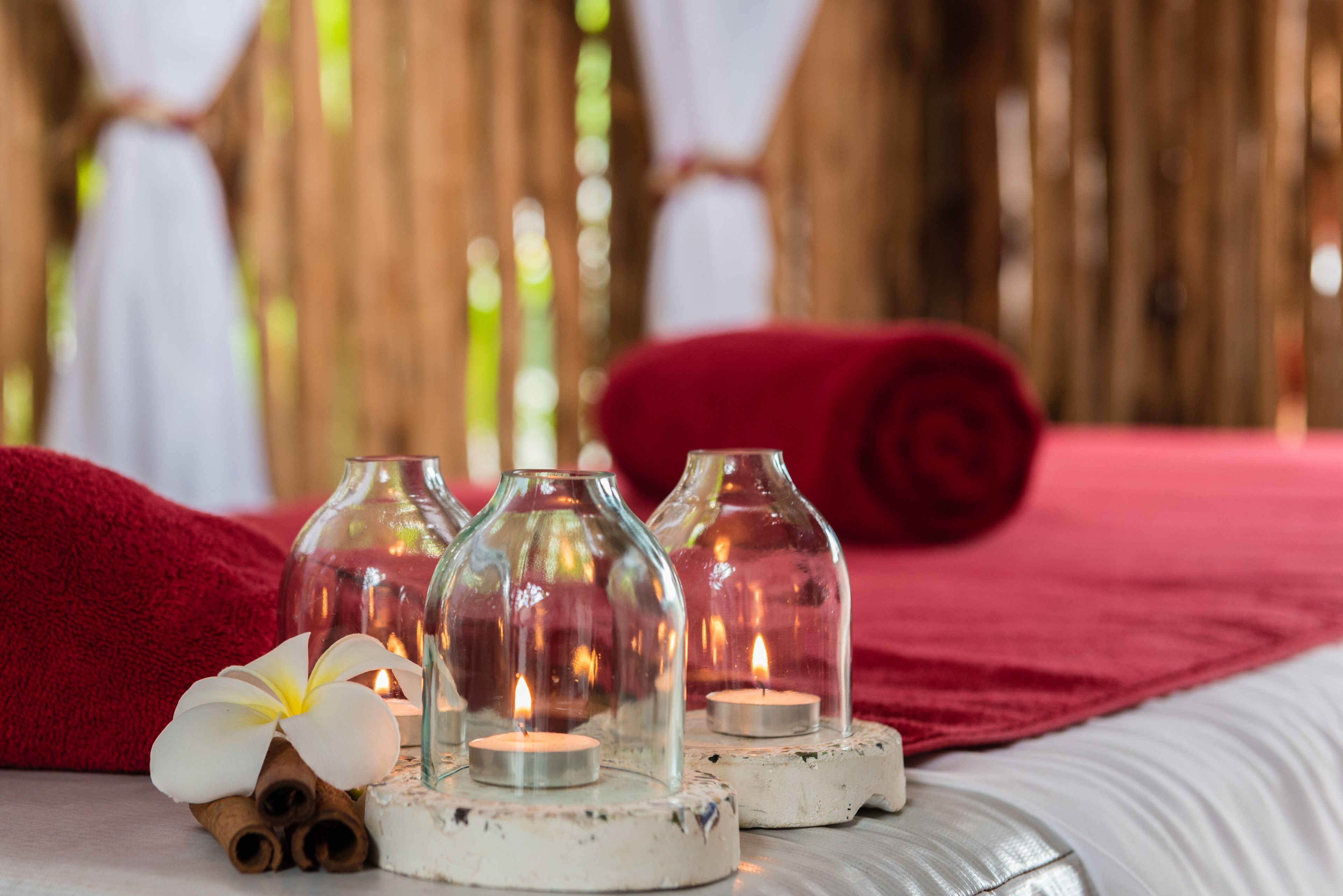A bed with red linens, a white pillow, and a red blanket. There are three glass candle holders with candles on a white coaster. A white flower and cinnamon sticks are on the bed.