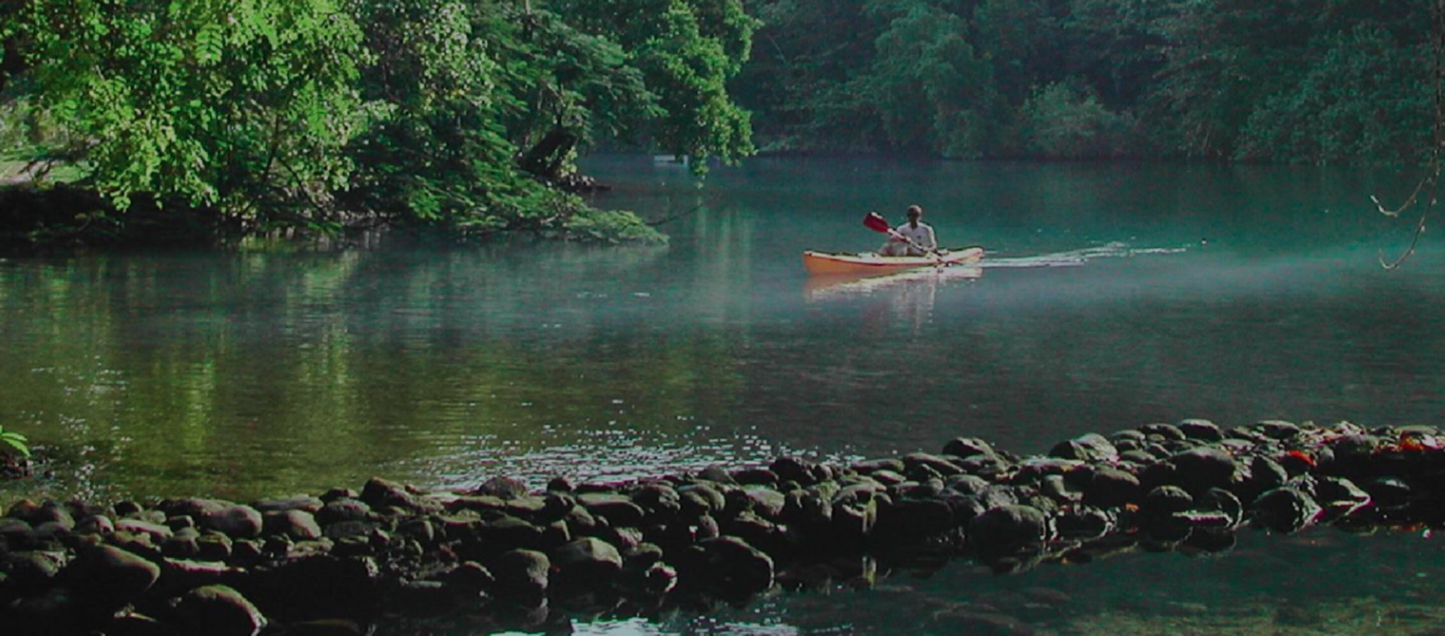 A person in a canoe on a calm river surrounded by rocks and dense greenery.