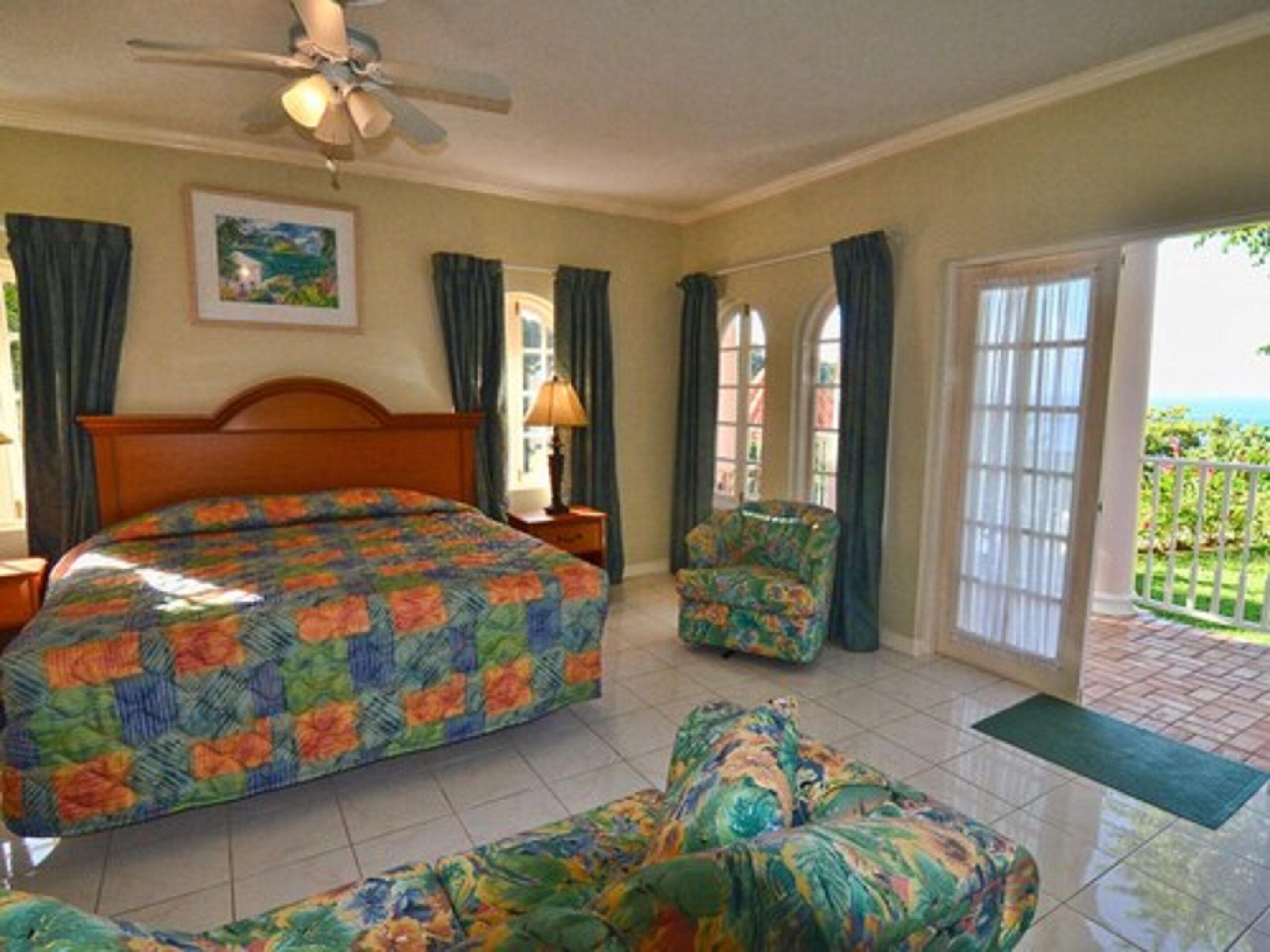 A bedroom with a bed, a chair, a nightstand, a lamp, a ceiling fan, and a view of the outdoors.