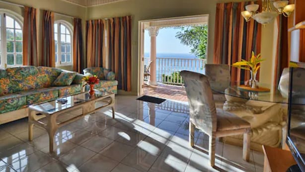 A spacious living room with a large sofa, a coffee table, and a chair. There is a view of the ocean through a glass door. - Tropical Lagoon Resort (Port Antonio)