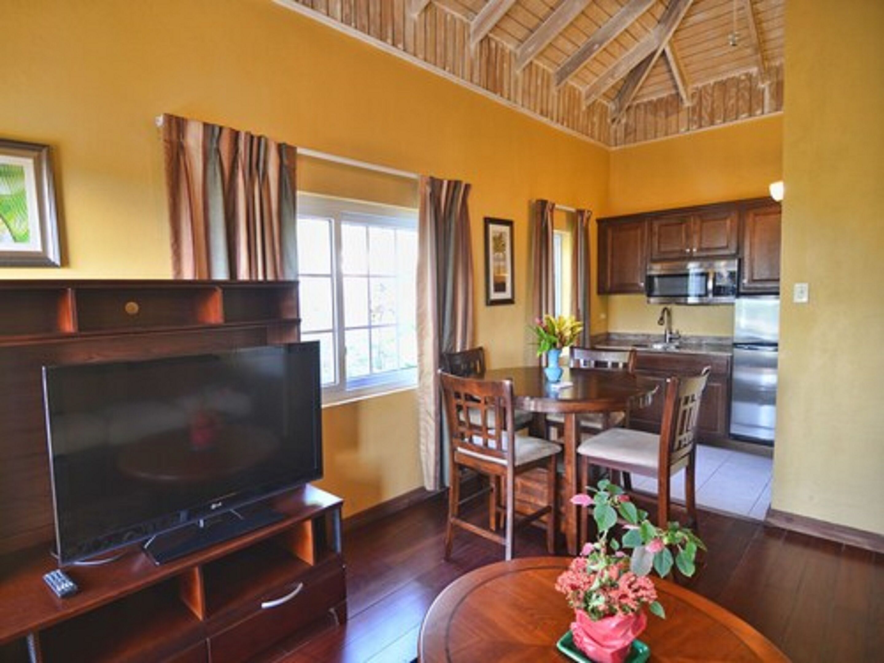 A living room with a TV, dining table, chairs, and a kitchen area with cabinets and appliances.