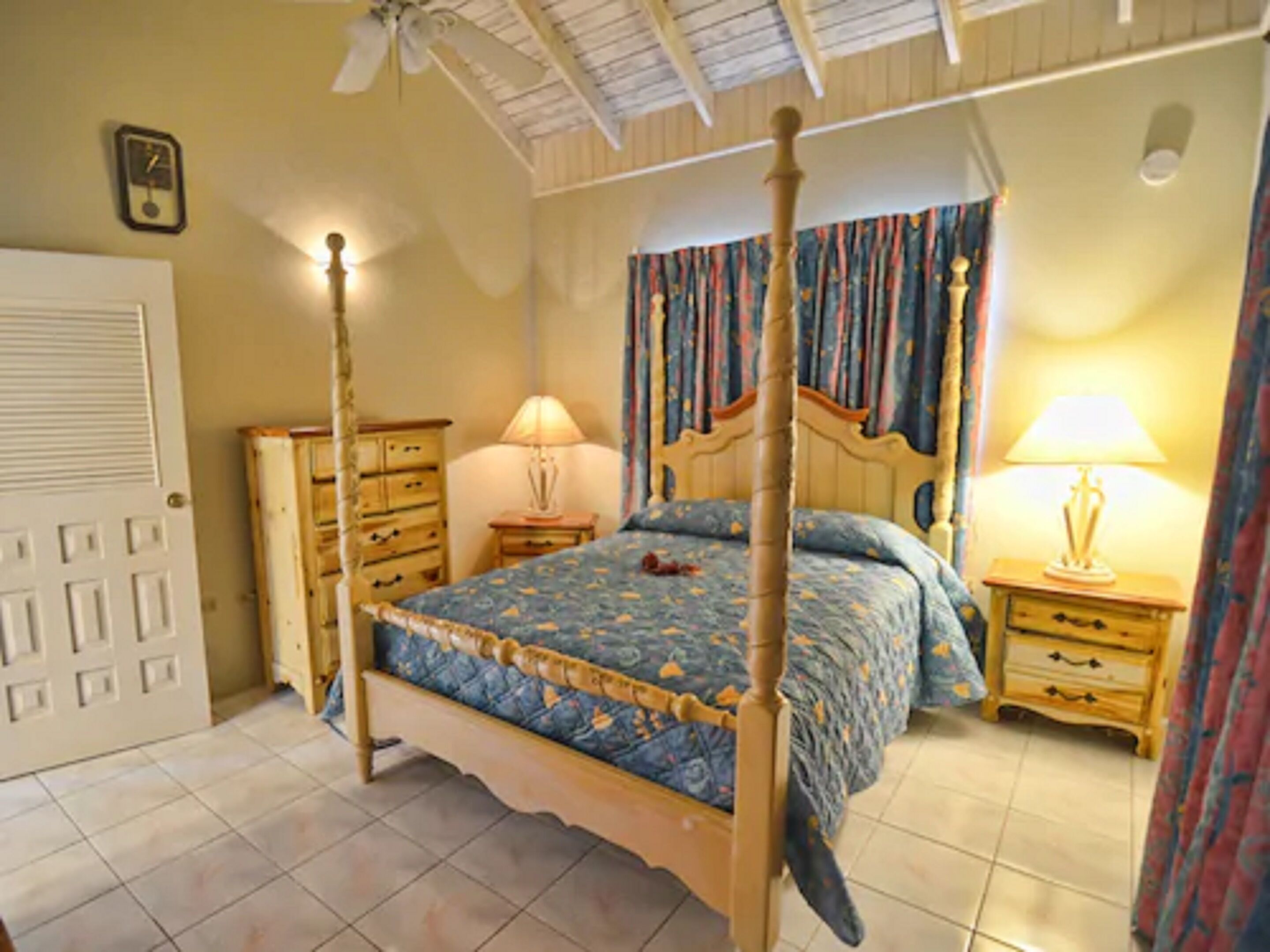 A four-poster bed with a floral bedspread, two bedside tables with lamps, a wooden dresser, and a ceiling fan.