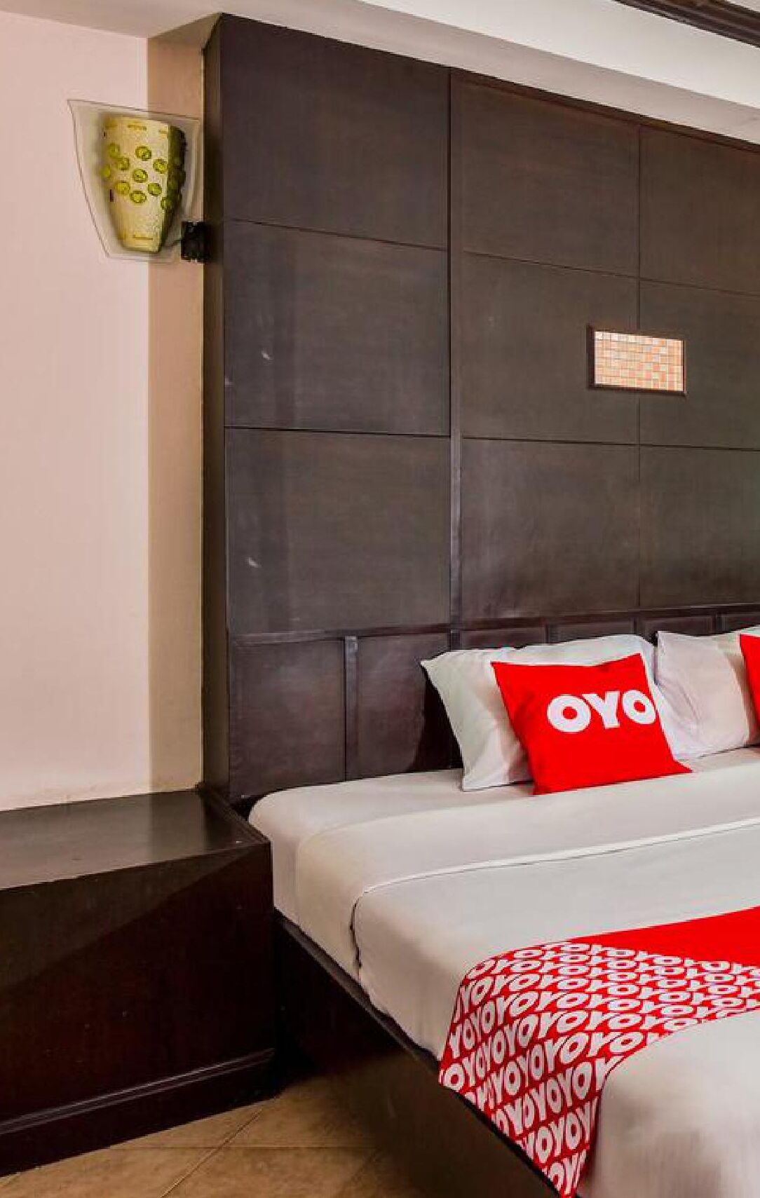 A hotel room with a dark headboard, a bed with white bedding and red pillows, and a small decorative item on the wall.