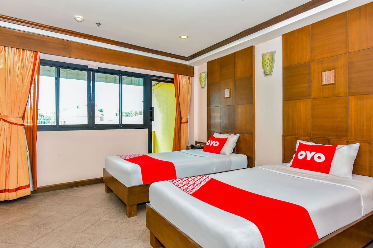 Habitación de hotel con dos camas, cada una con una colcha y almohadas rojas y blancas que tienen la palabra "OYO". La habitación tiene paneles de madera en las paredes y un ventanal con cortinas.