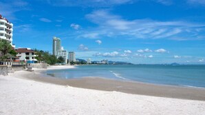 Una playa de arena blanca, cielo azul despejado y edificios al fondo.