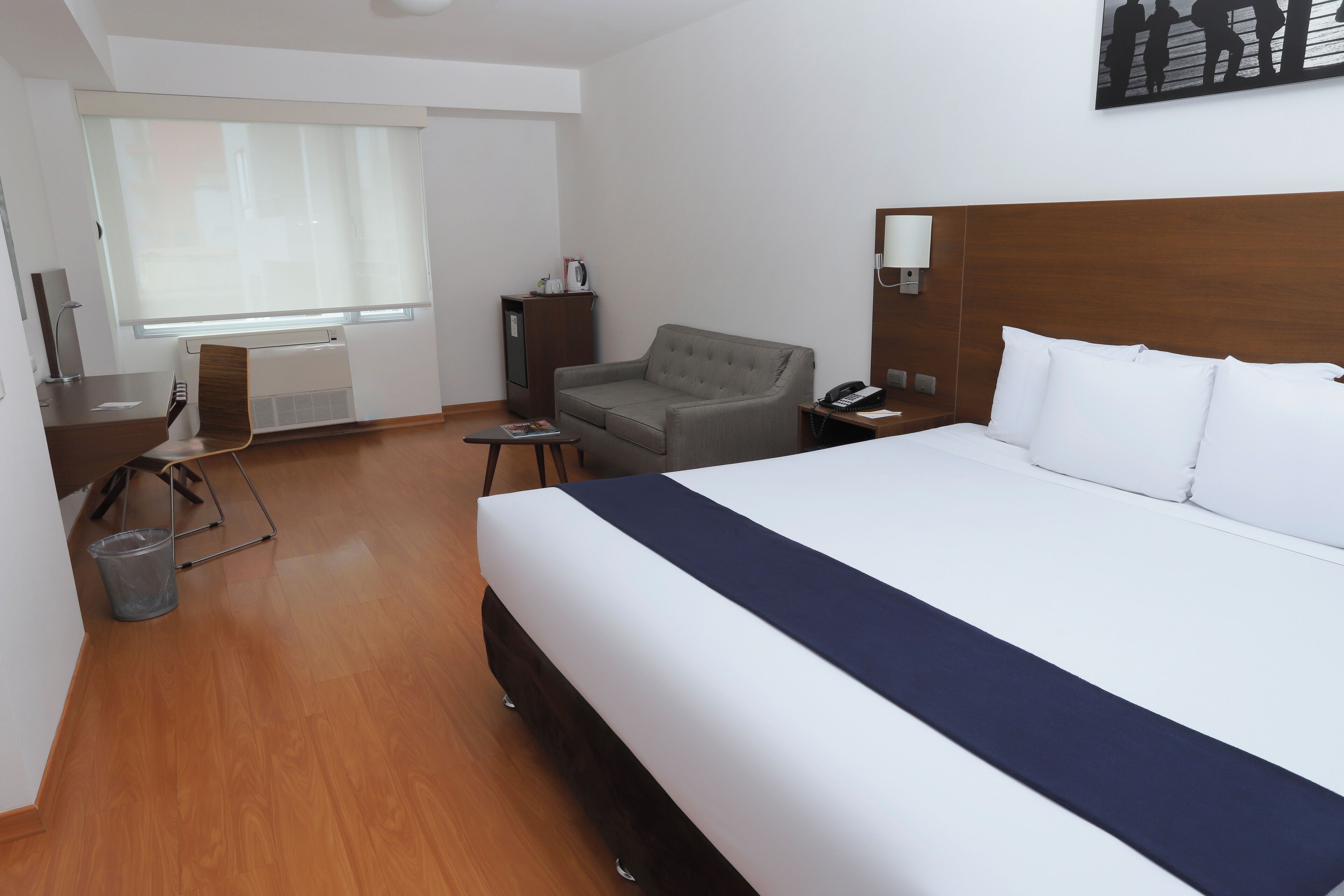 Una habitación de hotel con una cama grande, un sillón gris, un escritorio con silla, una mesita y una ventana con persianas.