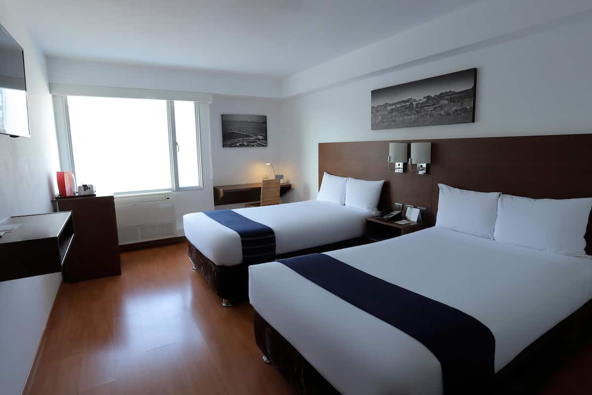 A hotel room with two beds, a desk, a chair, a nightstand, a lamp, and a window with a view.