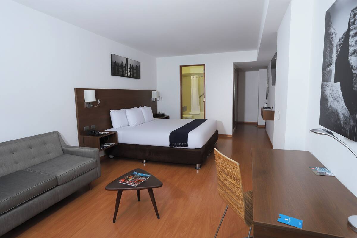 Una habitación de hotel con cama, sofá gris, una mesita y una silla.