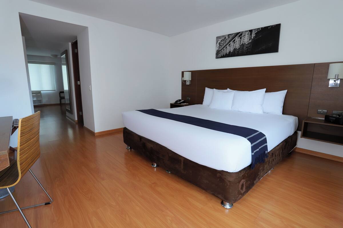 Una habitación de hotel con una cama grande, piso de madera, un escritorio con silla y una mesita de noche con lámpara.