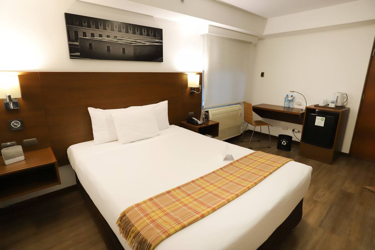 Habitación de hotel con una cama grande, un escritorio, una silla, una mesita de noche y una lámpara de pared.