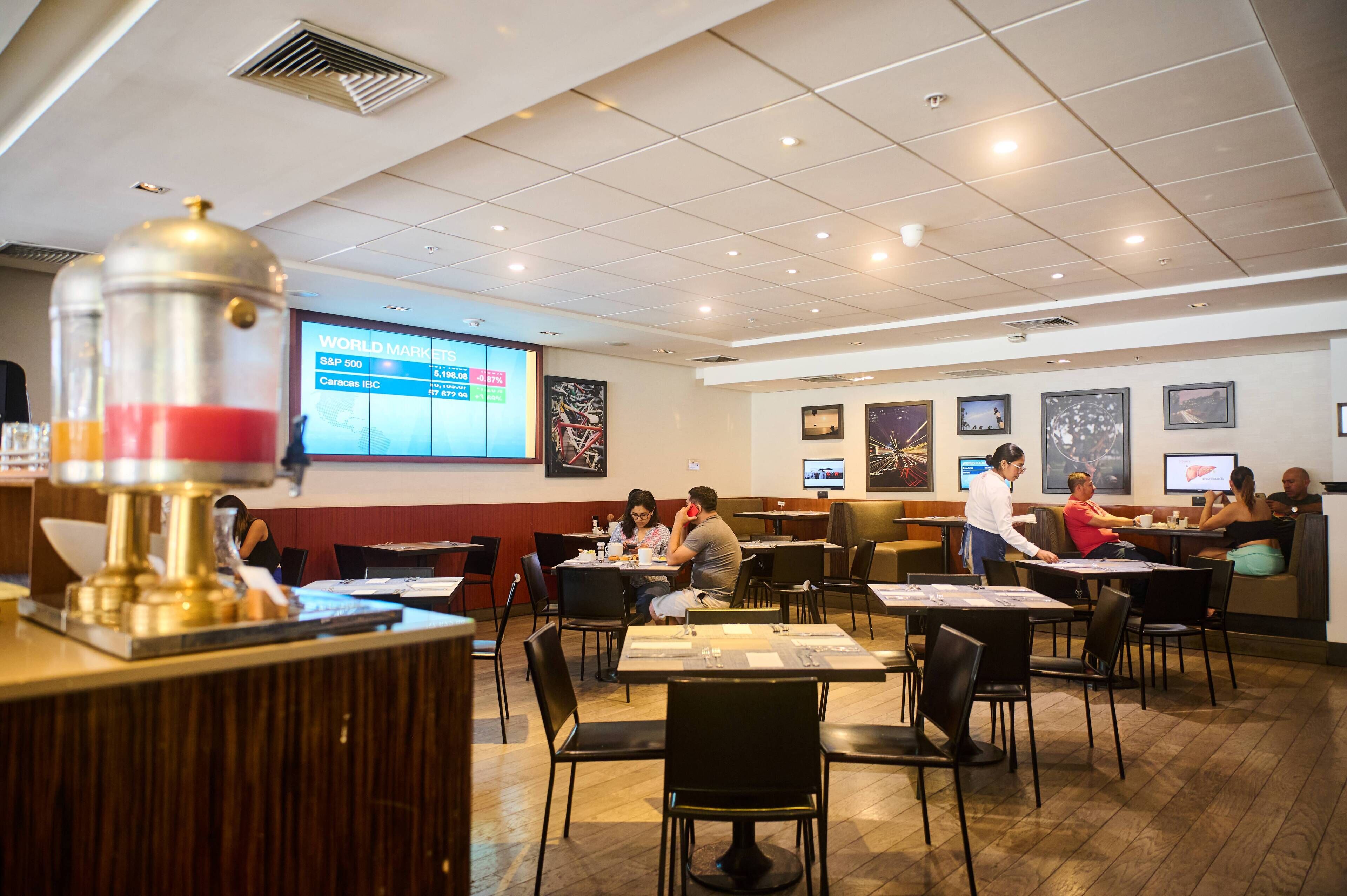 Un restaurante con mesas y sillas, una pantalla de proyección mostrando información del mercado de valores y una persona atendiendo en la barra.
