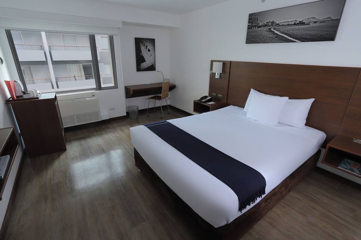 Habitación de hotel con una cama grande, un escritorio con silla, una mesita y una ventana con vistas a edificios.