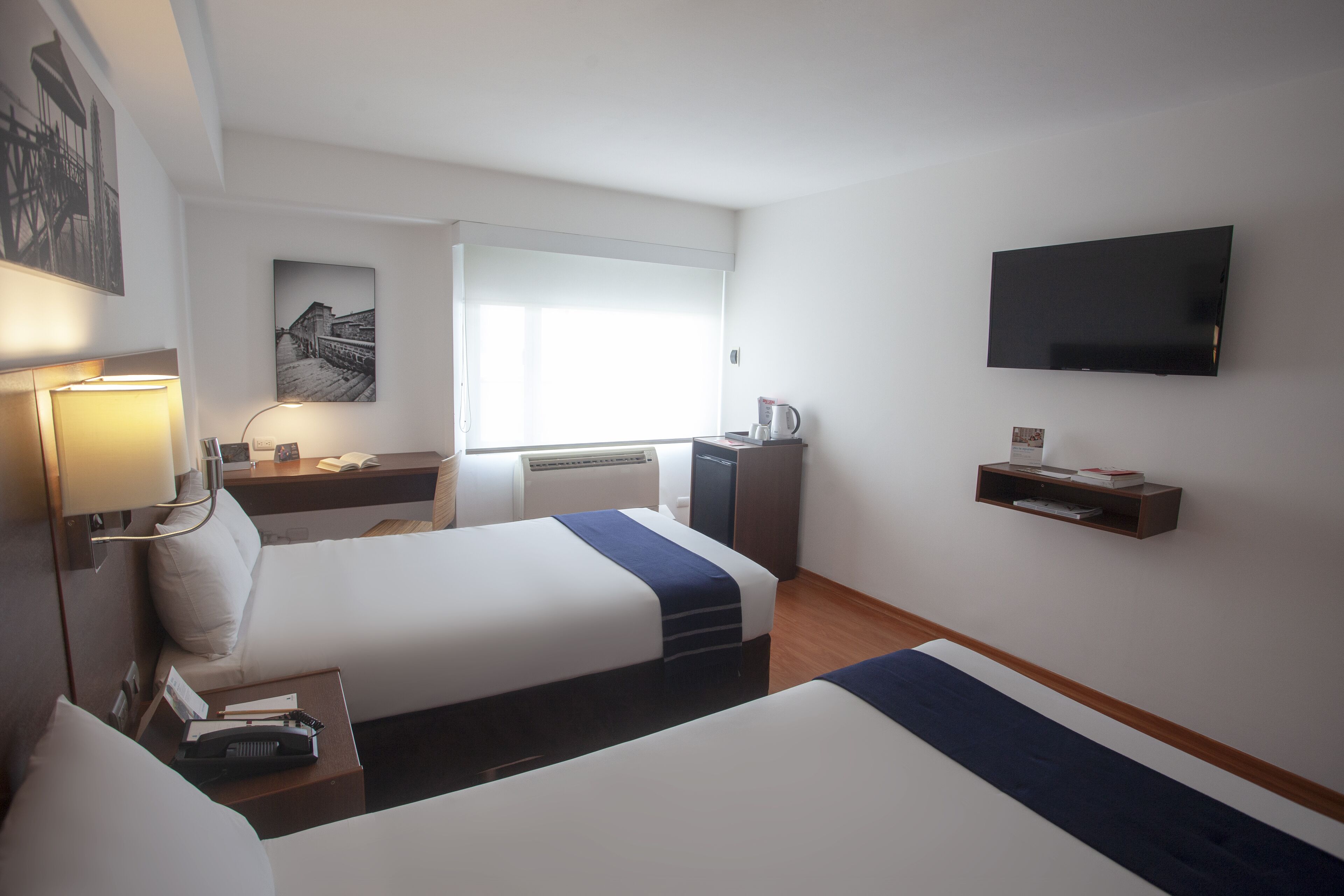 Habitación de hotel con dos camas, un escritorio, una silla, un televisor de pantalla plana y una ventana con persianas.