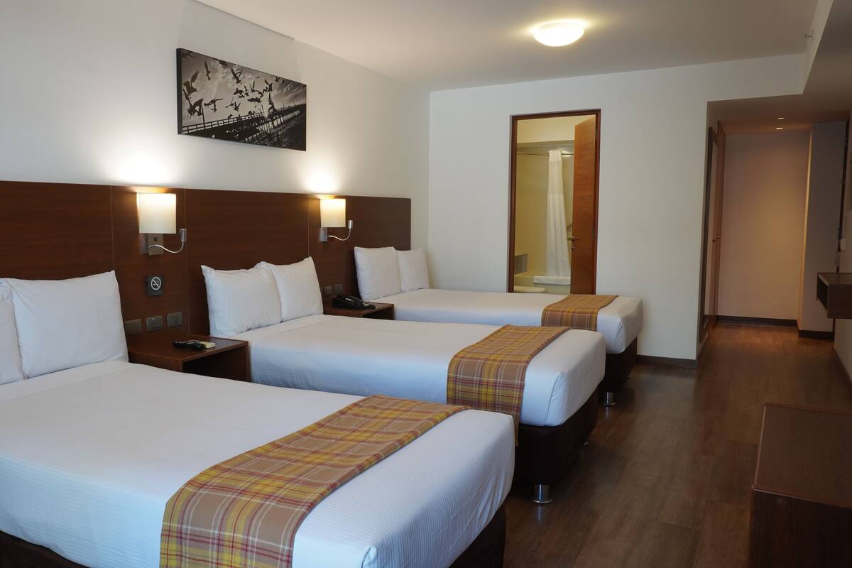 Habitación de hotel con tres camas, cada una con sábanas blancas y cabeceros de madera. Hay un cuadro en la pared sobre las camas, una mesita de noche con un teléfono y un espejo que refleja un baño con cortina de ducha.