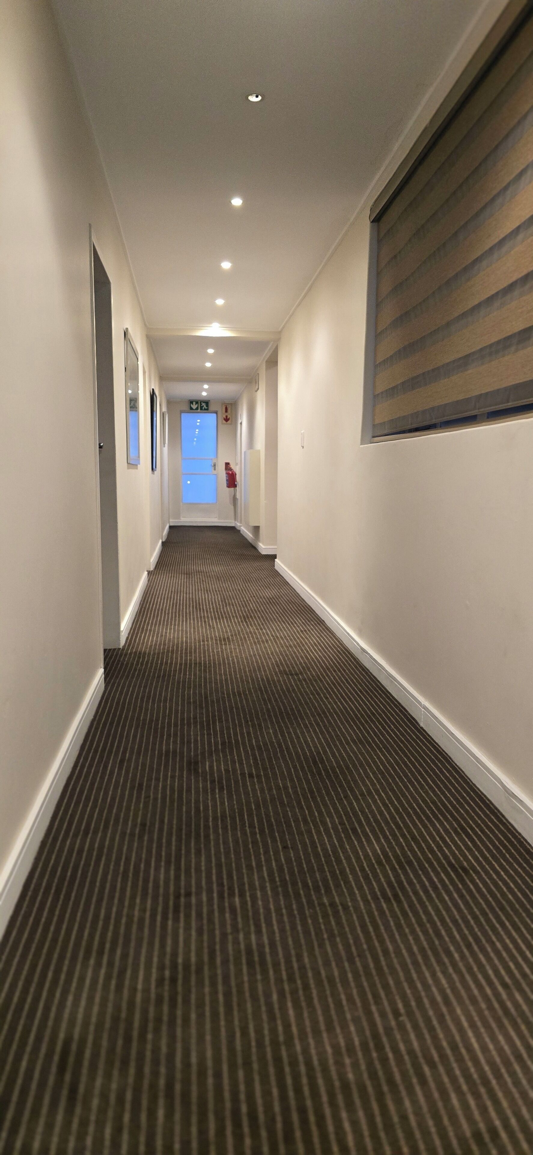 A hotel hallway with a striped carpet, white walls, and a window with blinds.