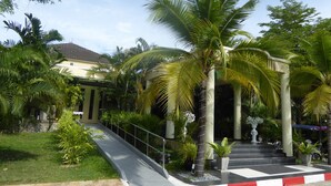 A tropical garden with palm trees, a paved walkway, and a building with a tiled roof. - Tropical Beach Resort Rayong (RAYONG)