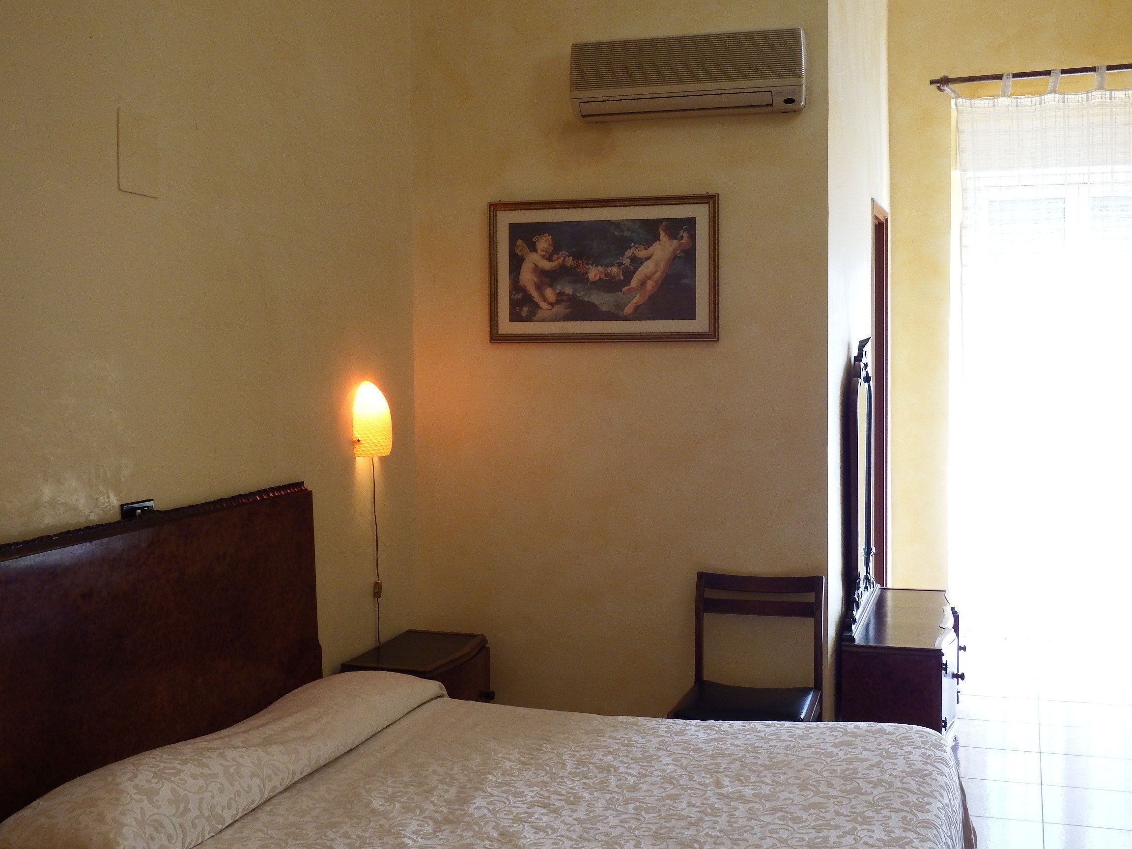A hotel room with a bed, a nightstand, a chair, a wall-mounted air conditioner, and a framed picture.