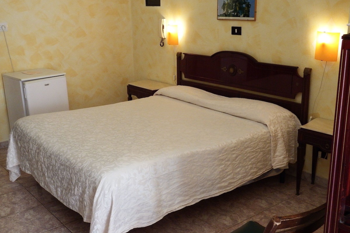 A neatly made bed with a white bedspread in a room with a small white refrigerator, a wooden nightstand with a lamp, and a wall-mounted light switch.